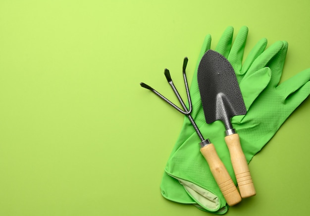 Фото Зеленые резиновые перчатки и садовый набор лопат, граблей, вил на зеленом фоне, вид сверху, плоская планировка
