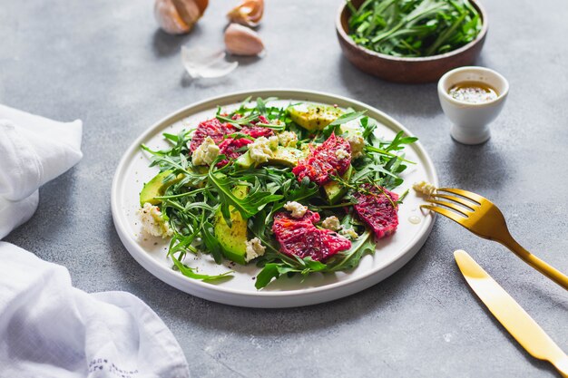 Arugula, 혈액 오렌지, 아보카도, 코 티 지 치즈 그린 포크 골드 나이프와 나이프, 화이트 린 넨 냅킨 블루 돌 벽에 그린 샐러드. 건강 한 먹는 개념. 평면도