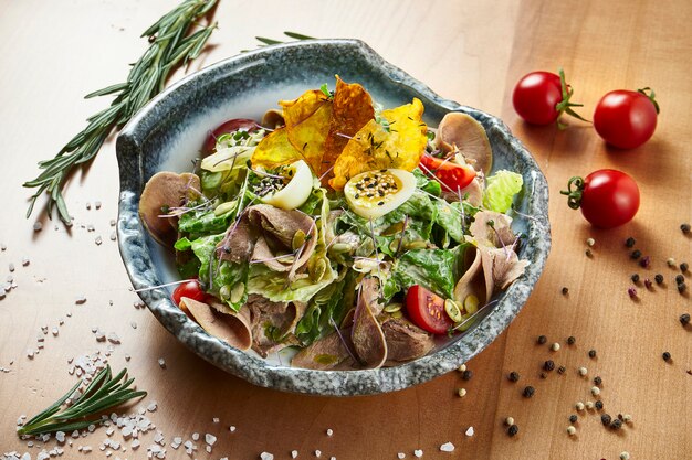 Green salad with lettuce, roasted veal tongue in a white bowl on a wooden surface in a composition with spices