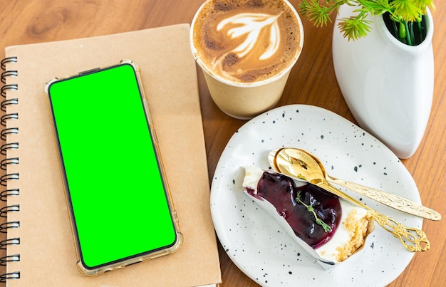 Зеленый экранкофе латте с латте-арт молочная пена в чашке из бумаги и смартфон в блокноте черничный торт на деревянном столе сверху как завтрак в кафе в кафе во время работы