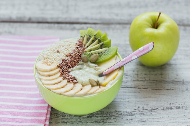 키위, 바나나, 사과, 건강한 채식주의 채식 아침 식사를위한 흰색 소박한 나무 배경에 씨앗을 얹은 녹색 스무디 그릇. 건강 식품 개념. 평면도