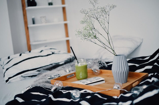 Green smoothie by flower vase in tray on decorated bed at home