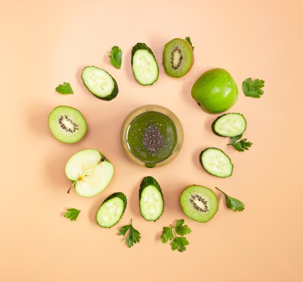 Зеленый коктейль в стеклянном стакане с семенами чиа на бежевом фоне. Нарезанные фрукты и овощи выкладываем по кругу. Здоровое питание, плоская планировка.