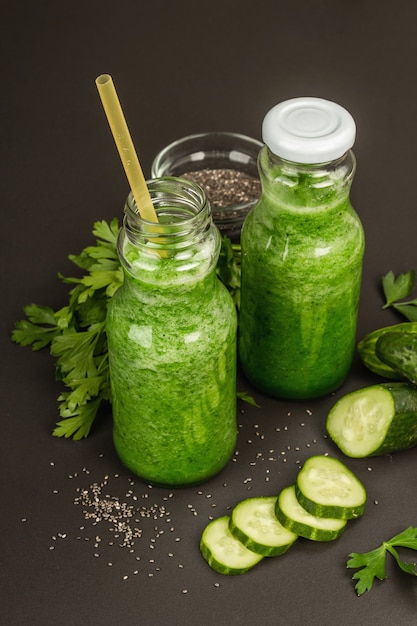 Green smoothie with cucumber in glass bottles. Fresh ripe vegetables, greens, and chia seeds. Black stone concrete background, close up