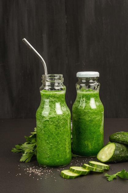 Green smoothie with cucumber in glass bottles. Fresh ripe vegetables, greens, and chia seeds. Black stone concrete background, close up