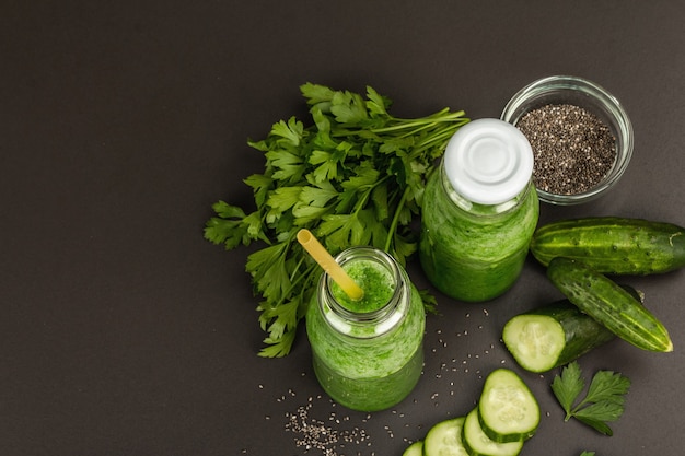 Green smoothie with cucumber in glass bottles. Fresh ripe vegetables, greens, and chia seeds. Black stone concrete background, top view