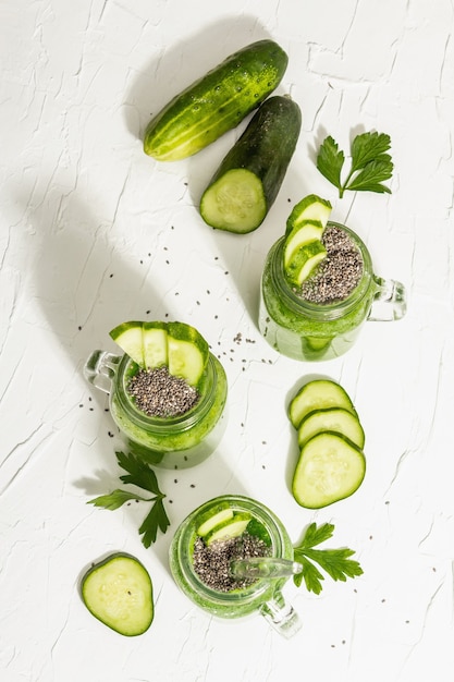 Green smoothie with cucumber in a glass jar. Fresh ripe vegetables, greens, and chia seeds. Trendy hard light, dark shadow. White putty background, top view