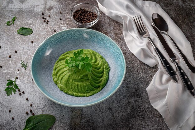 Green spinach and avocado cream soup in a blue plate with parsley and dill against a gray stone table