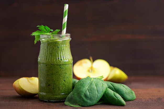 Green spinach smoothies in glass with ingredients on dark wooden table.
