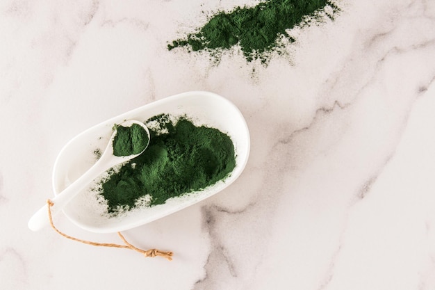 Green spirulina powder in an oval white bowl with a spoon on a marble background the concept of strengthening the body top view