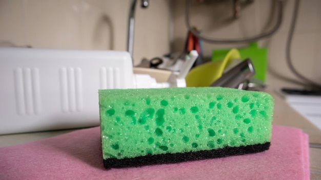 Green sponge and liquid soap dispenser for washing dishes on a dirty sink completely with dishes and kitchen utensils. Washing dishes in the kitchen by hand with detergent and a sponge.