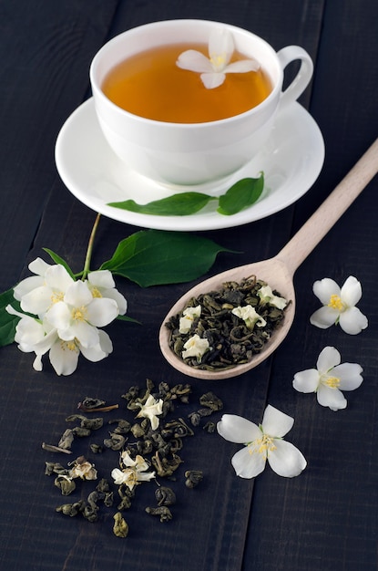 Tè verde con fiori di gelsomino e tazza di tè su fondo di legno scuro.