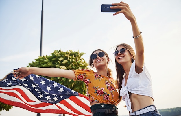 Green tree at background. Two patriotic cheerful women with USA flag in hands making selfie outdoors in park.