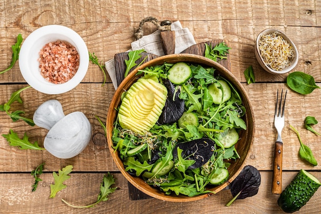 Green vegan salad with green leaves mix, avocado and vegetables. Wooden background. Top view.