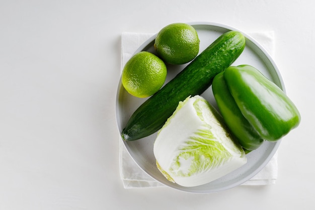 Зеленые овощи и фрукты на белой тарелке. Огурец, капуста, болгарский перец и лайм на белом столе. Вид сверху. Копировать пространство