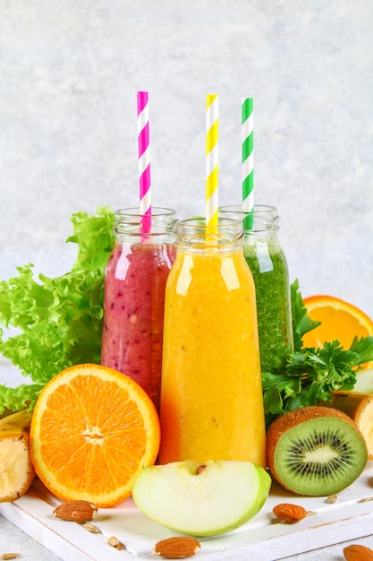 Green, yellow, purple smoothies in currant bottles, parsley, apple, kiwi, orange on a gray table