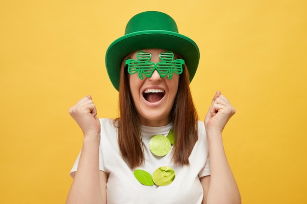 Photo greenthemed event irish culture celebration overjoyed caucasian woman wearing green leprechaun hat and clover glasses standing isolated over yellow background with clenched fists