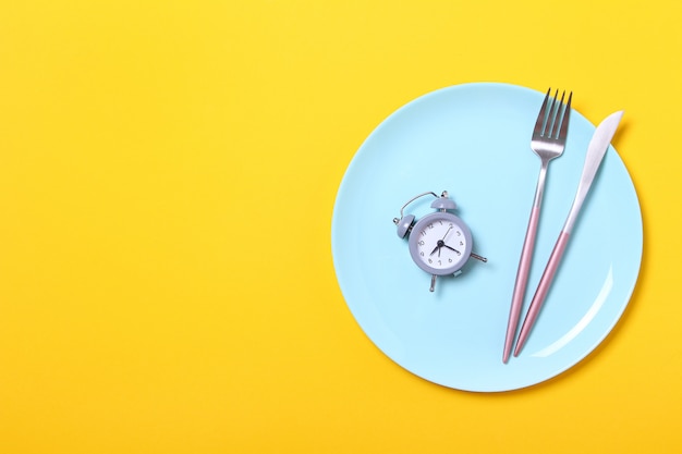写真 灰色の目覚まし時計、フォーク、黄色の空の青いプレートにナイフ。断続的な断食、ランチタイム、ダイエット、減量のコンセプト。