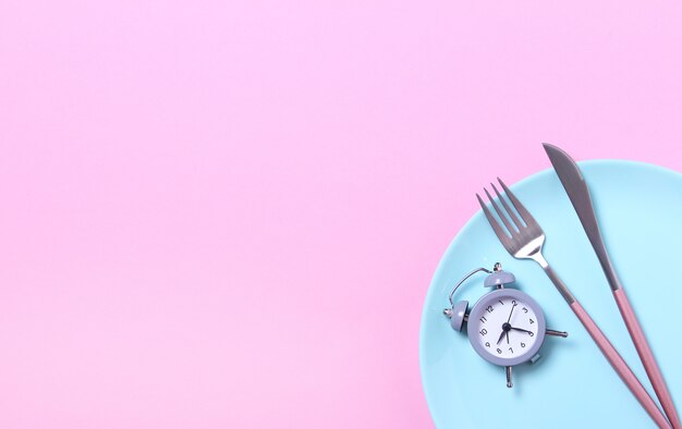 Foto sveglia, forchetta e coltello grigi in piatto blu vuoto sul rosa concetto di digiuno, ora di pranzo, dieta e perdita di peso intermittenti vista superiore, disposizione piana, minimalismo.