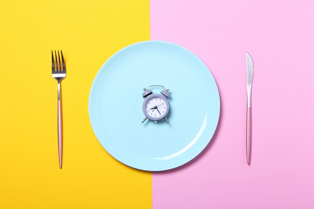 Grey alarm clock,fork and knife in empty blue plate on yellow and pink .Concept of intermittent fasting, lunchtime, diet and weight loss.Top view,flat lay,minimalism.