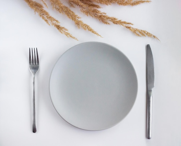 Grey empty plate with silver cutlery on the white table decorated by dried flowers