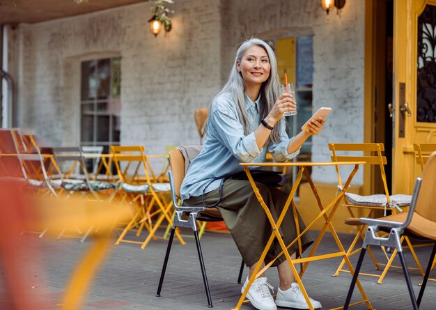 야외 카페 테라스에서 유리와 스마트폰을 가진 회색 머리 아시아 여성