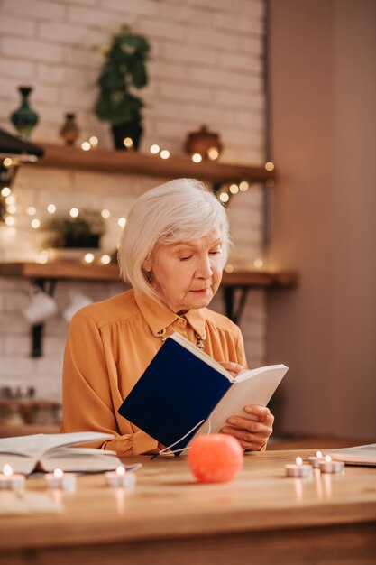 Grey-haired beautiful elderly lady in orange blouse reading a book