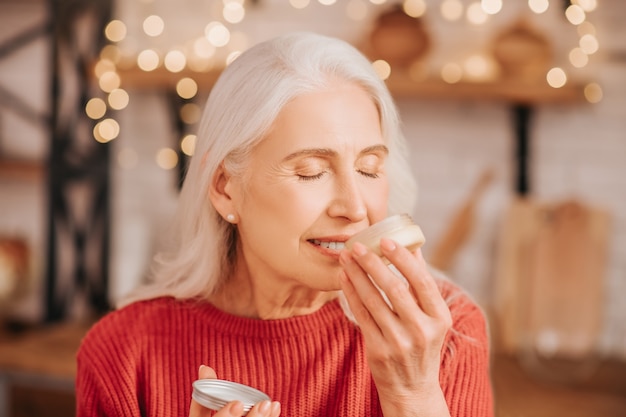 Grey-haired beautiful lady in red smelling new cream