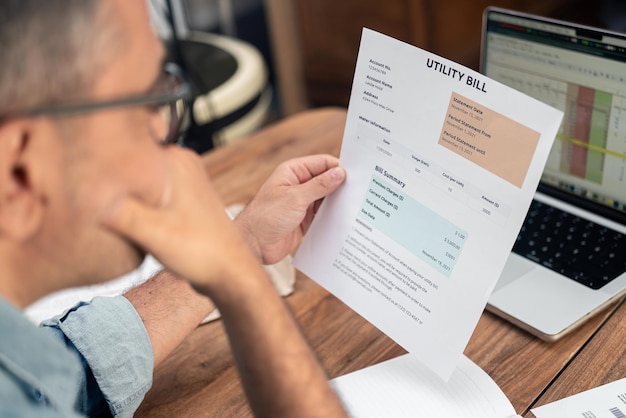Grey haired male employee looks at utility bill attentively sitting at office workplace financial