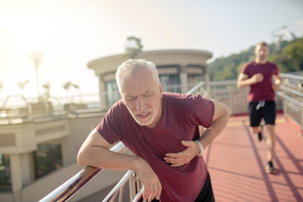 Grey-haired male frowning, holding his hand on his chest, young male jogging behind him