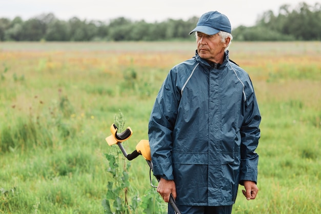 Grey haired mature man posing outdoor, holding metal detector in hands, standing in field looking away with pensive expression