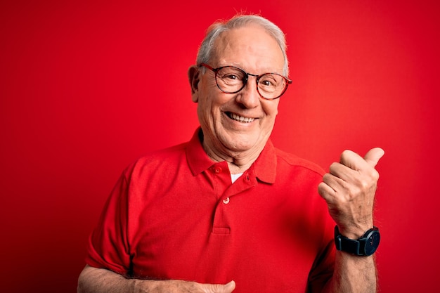 Photo grey haired senior man wearing glasses and casual tshirt over red background smiling with happy face looking and pointing to the side with thumb up