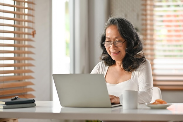 Grey haired senior woman in glasses working remotely from home office with laptop computer