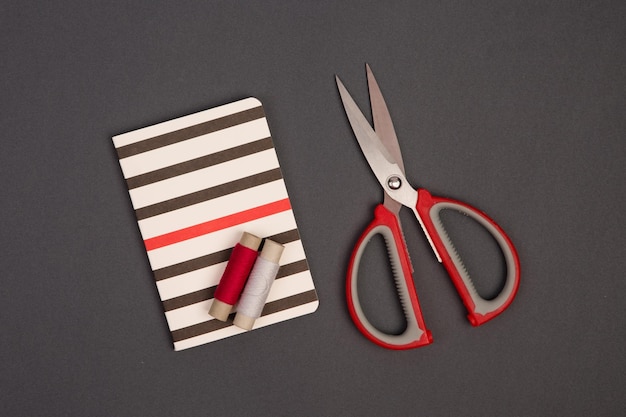 Grey and red thread scissors and striped notepad for notes on\
grey background