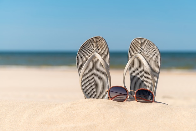 Grey sandals at the beach on a beautiful sunny day. Slippers in the sand by the sea. Flip flops at the shore by the ocean.