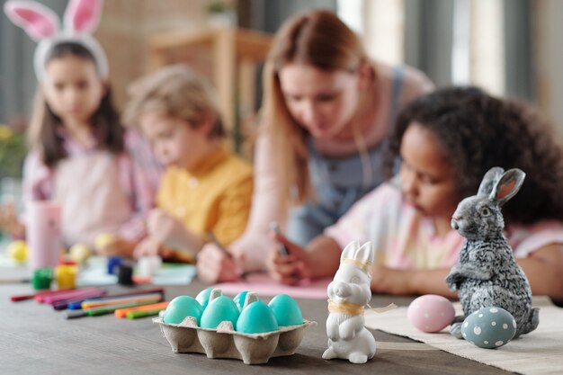 Grey and white toy rabbits and group of painted Easter eggs in egg-box standing on wooden table with kids drawing pictures on background