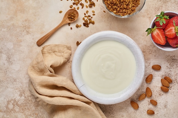 Griekse yoghurt in witte kom met ingrediënten voor het maken van ontbijtgranola en verse aardbeien op oude beige lijst. Bovenaanzicht.