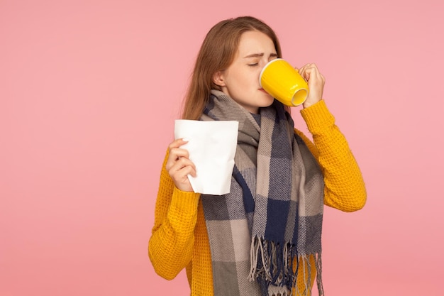 Griep behandeling. Portret van een ziek gembermeisje dat een grote sjaal draagt, een servet vasthoudt en hete thee drinkt, griep en hoge temperaturen behandelt, gezondheidszorg. indoor studio-opname geïsoleerd op roze achtergrond