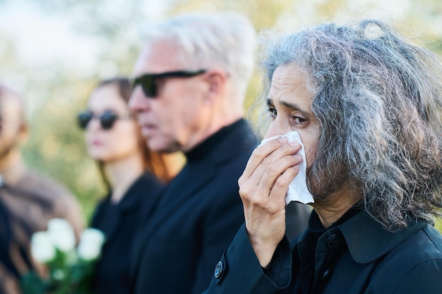 Grieving senior woman wiping tears by handkerchief while crying at funeral