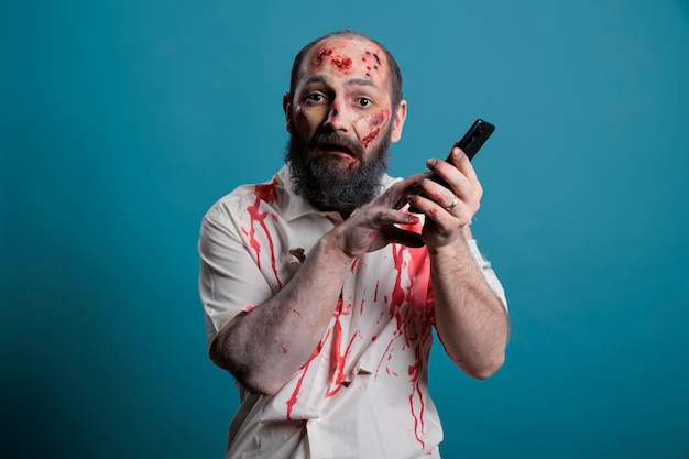 Griezelige duivel die smartphone in studio vasthoudt, app voor mobiele telefoons gebruikt terwijl hij agressief en eng is. Angstaanjagende ondode zombie met bloedige wonden, wreed kwaad lijk met telefoon.