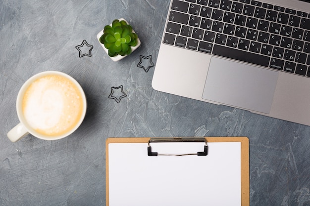 Grijs bureau met zilveren laptopcomputer en blanco papieren lijst, koffiekopje en plant