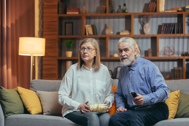 Grijsharig ouder echtpaar thuis kiest een televisieprogramma om naar te kijken en schakelt van kanaal met een tv-afstandsbediening Ouder volwassen gezin man en vrouw kunnen geen programma kiezen om nieuws of film te kijken