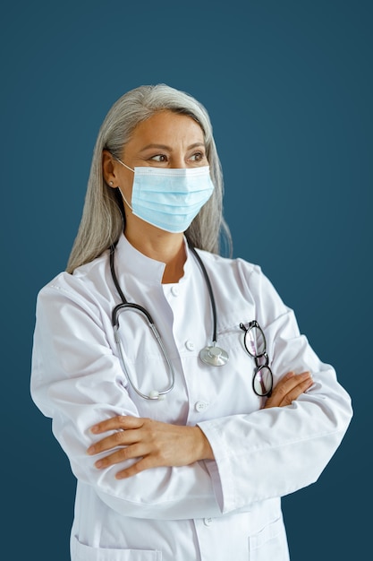 Grijsharige Aziatische vrouw arts met masker en gekruiste armen staat op blauwe achtergrond