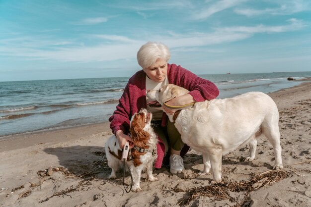 Grijsharige dame zit op haar hurken op het zandstrand en knuffelt haar twee honden