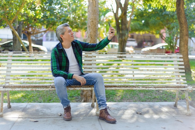 Grijsharige man zit op een bankje in het park en neemt een selfie