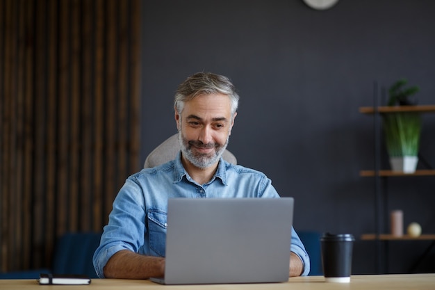 Grijsharige senior man aan het werk in kantoor aan huis met laptop. Zakelijk portret van knappe manager zittend op de werkplek. Online studeren, online cursussen. Bedrijfsconcept.