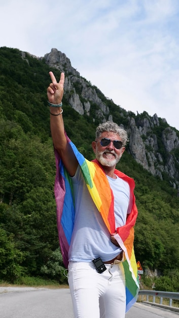 Grijsharige senior oudere blanke man biseksualiteit diabeticus met een baard en zonnebril met een regenboog LGBTQIA-vredesvlag in bergen Viert Pride Month Rainbow Flag Day gay parade Verticaal