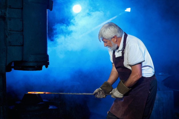 Grijsharige smid aan het werk met gesmolten metaal bij smidse