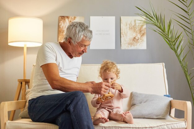Foto grijsharige volwassen man met wit t-shirt zittend op de bank met zijn babykleindochter in de huiskamer spelen met eco houten speelgoed samen genieten van vrije tijd