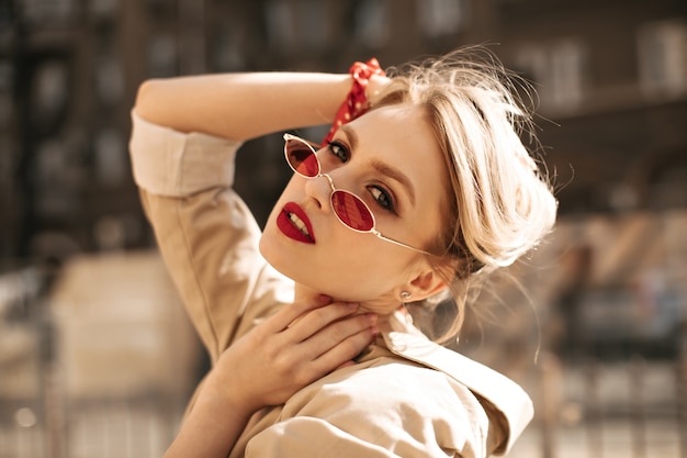 Grijze-eyed blonde vrouw met rode lippen in kleurrijke zonnebril en beige trenchcoat kijkt in de camera en raakt haar buiten aan